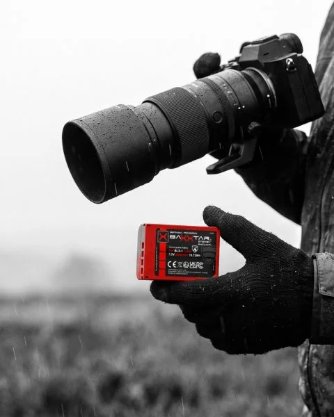 Naturfotograf im Regen hält einen roten Baxxtar BLX-1-Akku für OM System / Olympus