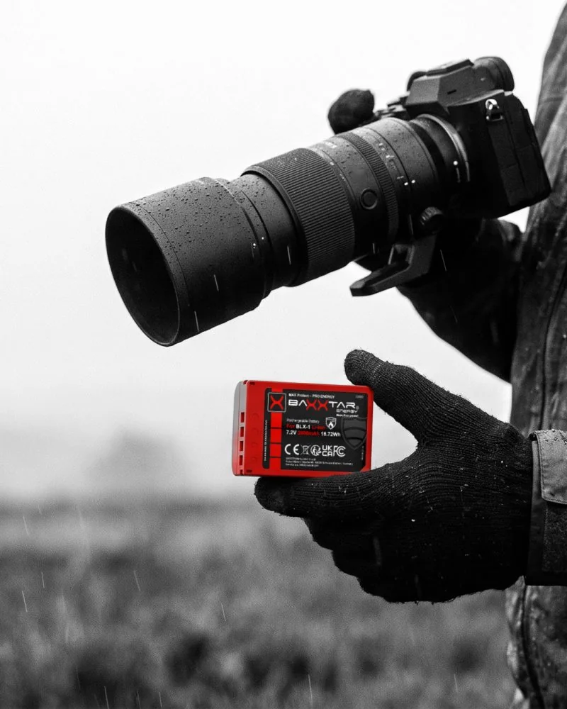 Naturfotograf im Regen hält einen roten Baxxtar BLX-1-Akku für OM System / Olympus